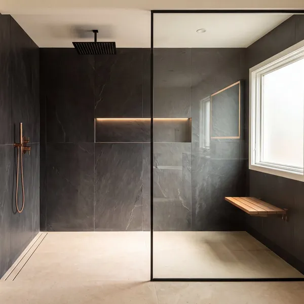 A curbless walk-in shower in dark slate with a frameless glass panel, rain head, lit niche and teak bench