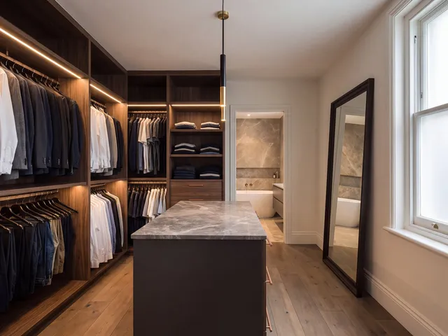 A walk-in dressing room with walnut wardrobe joinery, integrated shelf lighting and an island dresser, opening to the ensuite
